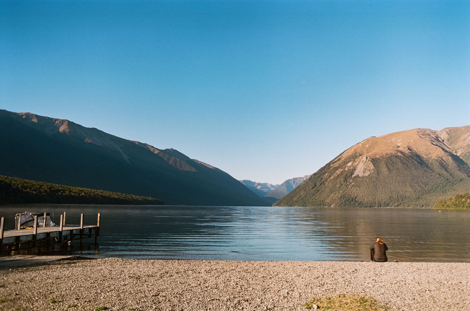 A girl sits, back turned to the camera, on the shore of a glacial lake, looking out into the mountains that frame the lake in the horizon. A dock extends a few meters into the lake on the left.